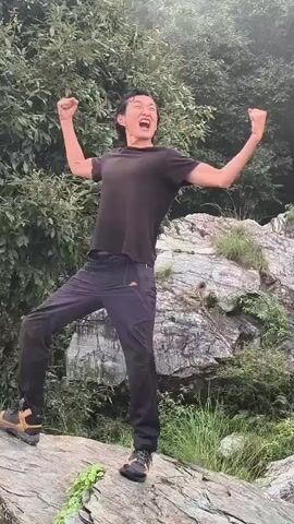A person is standing on a rock, celebrating with their arms raised in the air. They are wearing dark athletic pants and a dark t-shirt. The background is a lush green forest with large rocks and trees. The sky is overcast, and it appears to be misty or foggy.