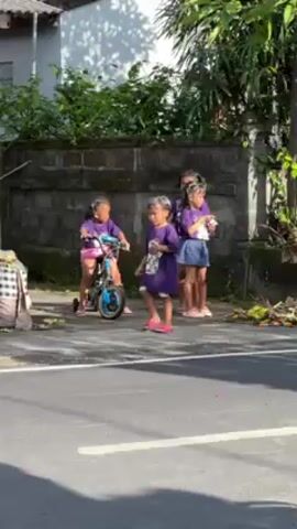 In a sunny outdoor shot, four young girls are seen near a road and a gray stone wall, flanked by lush green trees. The girls are dressed in matching purple shirts with an image printed on the front. One of them is walking alongside a small blue bicycle, while the other three stand together, holding hands. The girl with the bicycle wears pink sandals and has white paint or cream on her hair. Two of the girls wear denim shorts, while the third wears a denim skirt. The scene includes a section of a house with a gray tiled roof, and the road shows signs of some scattered debris. A checkered cloth-covered object sits on the side of the road next to the girls. The group's interaction suggests a casual, everyday scene.