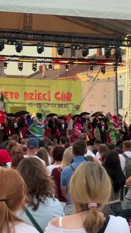 A wide shot shows an outdoor stage where a group of performers in colorful, traditional Mexican attire, including sombreros and ruffled dresses, are dancing. A banner behind them reads "ŚWIĘTO DZIECI GÓR 19-25 LIPCA 2026". A crowd of spectators, with their backs to the camera, is watching the performance. The lighting suggests it is late afternoon or early evening.