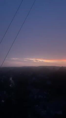 A time-lapse video shows a twilight sky with a gradient of deep blue at the top fading to shades of purple and pink near the horizon. A thin, bright orange band of light is visible where the sky meets the horizon, suggesting the last rays of a sunset. Below the sky, a silhouetted landscape of rolling hills and trees is visible, dotted with the dark shapes of houses and structures. In the foreground, dark wires stretch diagonally across the frame from the top left. The scene is dimly lit, with the details of the landscape obscured by shadow.