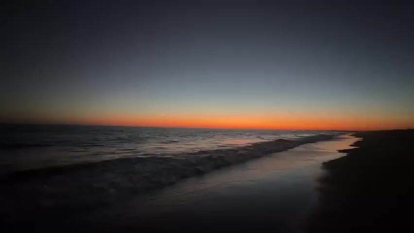 The video shows a wide shot of a beach at dusk. The sky is a gradient of dark blue at the top, transitioning to a vibrant orange near the horizon, indicating the sun has recently set. The ocean is visible on the left side of the frame, with gentle waves rolling towards the shore. The water reflects the orange and blue hues of the sky. On the right side of the frame is the dark silhouette of the beach. The overall scene is peaceful and serene. There are no people or objects clearly visible on the beach. The video is static, with no camera movement.