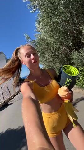 A young woman with blonde hair pulled back in a ponytail, wearing a yellow sports bra and shorts, holds a yoga mat, a water bottle, and a banana. She is walking outdoors on a paved path, with trees and buildings in the background under a clear blue sky. The camera is held at a low angle, suggesting it is being used for a selfie or vlogging.