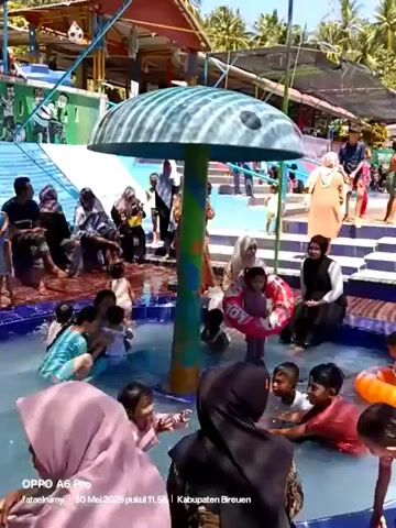 The video shows a group of people, mostly children and women wearing hijabs, enjoying a sunny day at a water park. The children are playing in a shallow pool, some with inflatable rings, under a large, mushroom-shaped water feature. Adults are seated around the pool’s edge, some watching the children. The backdrop includes other water park structures, greenery, and people on higher levels. The video seems to be taken on a mobile phone, as indicated by the watermark at the bottom that contains the phone model and location.
