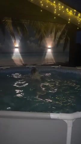 A young girl swims in a small above-ground pool at night. She is wearing a bikini and is facing the camera. The pool is lit by overhead string lights and two spotlights, which shine through artificial palm trees onto a beach scene painted on the wall behind the pool. The water is agitated and appears to have jets or bubbles. The pool has a white frame.