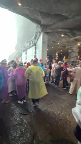 A crowd of people wearing rain ponchos in various colors stand inside a cave or tunnel, many holding up their phones to take pictures or videos. The opening of the cave frames a bright, hazy view of a waterfall cascading between two cliffs. The atmosphere appears damp and misty, with water visible on the ground. The people seem to be admiring and recording the natural wonder of the waterfall.
