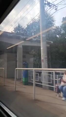 The video shows a view from a moving train or vehicle. The camera is pointed outwards, capturing a platform or walkway area. The scene is dominated by a concrete canopy with overhead lighting. To the right, there is a white brick building with windows. Further down the platform, people are visible, some with luggage. There are also metal railings and signage. Beyond the platform, trees and foliage are visible, suggesting an outdoor environment. The sun is shining, casting light and shadows. The camera motion creates a sense of movement and passing scenery.