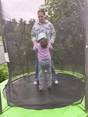 A video shows a young girl and a woman jumping on a trampoline in a backyard. The woman has dark hair pulled back, is wearing a light blue and white striped long-sleeved shirt and blue jeans, and is holding the girl's hands. The girl has brown curly hair, is wearing a light blue hat, a pink and white striped shirt, and blue jeans. The trampoline has a black jumping mat, a green and black frame, and a black safety net around it. The background shows green trees and a fence. The video ends with the woman holding the girl's hands while the girl jumps.