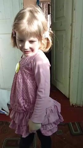 A young girl with blonde hair in pigtails wearing a pink dress with a smiley face pin twirls in front of an open doorway. The girl stops twirling and smiles at the camera. A small amount of debris is scattered around the floor around the doorway. A plastic bag is on the floor nearby. A glimpse of a room with curtains can be seen through the open doorway.