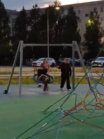 Two young people are playing on a playground. One person is swinging on a circular swing, and another person is pushing the swing. In the background, there is a large apartment building, trees, and cars parked on the street. The playground has a green rubber surface and a large climbing structure made of ropes and poles. The lighting suggests it is dusk or early evening.