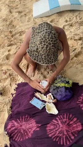 A fair-skinned person in a leopard print hat crouches on a purple and pink tie-dye blanket on the sand. The person's back is to the camera, and they are seen holding a cup with a lid. The blanket is spread on the sand. Other items on the blanket include a light blue iPhone, three large seashells, and a geometric-shaped object that is purple and green. Behind the person is a white wicker chair with blue stripes.