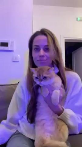 A woman with shoulder-length light brown hair is holding a fluffy, orange-colored cat. She is wearing a white sweater. The cat looks sleepy and is being held upright, its paws slightly dangling. They are both seated on a taupe-colored couch. Behind them, the wall is white, and there is a small white electronic device mounted on the wall, along with a light switch. There is a doorway to another room visible to the right. The lighting is bluish. The woman makes a kissing face at the camera. Music is playing in the background.