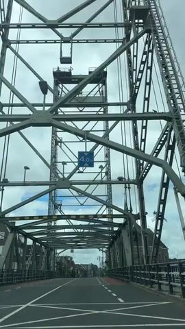 The video shows a view from inside a vehicle moving across a large metal bridge. The camera is pointed forward, capturing the intricate steel structure of the bridge overhead. A blue sign with "4.0m" is visible, indicating a height restriction. The road ahead has lane markings and a pedestrian walkway with cyclists. Buildings line the street beyond the bridge. The sky is cloudy. The vehicle appears to be driving on the right side of the road.