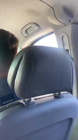 The video is taken from the back seat of a car, looking forward. The camera is focused on the back of the driver's head and shoulders. The driver is wearing a dark blue collared shirt with a repeating pattern of small interlocking shapes. On the left side of the driver's shirt, there is a dark rectangular patch with white text, partially obscured by the seat headrest. The car's dashboard is visible in front of the driver, including the car's radio display, which shows a music player interface. Above the dashboard, a rearview mirror is visible, with a car air freshener hanging from it. The windshield shows a road with other cars, buildings, and trees. The sky is bright and appears to be daytime. The car appears to be in motion, driving down a paved road. There are traffic signals and road signs visible outside the car. The interior of the car has a sunroof.