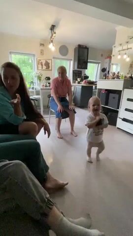 In a bright, well-lit room, a toddler with light brown hair stands in the center, wearing a white t-shirt and light-colored leggings. The toddler is holding a dark-colored object, possibly a phone, and appears to be looking at it or interacting with it. The toddler is smiling and facing slightly to the left. In the background, a woman with blonde hair sits on a chair, wearing a pink shirt and blue skirt. She is looking towards the toddler. To the left of the frame, two people are partially visible, seated on a couch. One person, with long brown hair, is on the far left, and another person is in the foreground, wearing gray pants and white socks. The room has a wooden floor and appears to be a living area or kitchen, with furniture and decorations visible. The overall atmosphere seems relaxed and familial.