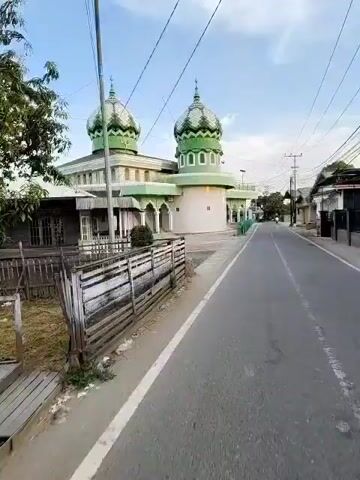 The video shows a street view in front of a mosque. The mosque has two green domes and a light-colored minaret. The building is painted in green and white. A road runs alongside the mosque, with a wooden fence on the left side and a green metal fence in front of the mosque. The sky is blue with some clouds. The scene appears to be in a residential area, with houses visible in the background. The video is taken from a moving vehicle, suggesting it is a walk-through or drive-by.