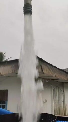 A long pipe expels a forceful jet of water downwards. The water appears opaque white and is turbulent. The pipe is held against a cloudy sky, with its textured surface suggesting it's made of metal and rubber. Below the pipe, a small portion of a building is visible. The building has a corrugated metal roof with some damage. The wall is white, and there's a wooden door with vertical planks on the right. A blue pipe is partially visible in the lower left corner. The overall environment suggests an outdoor setting, possibly a rural or industrial area.