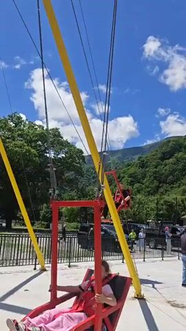 The video shows a person (a man) riding a giant swing set outdoors. The swing is red and features a reclining seat. The structure supporting the swings consists of large red and yellow poles and cables.

The scene is set alongside a wide, fast-flowing river with rapids. Across the river, lush green mountains covered in trees are visible under a clear blue sky with scattered white clouds. The person swings out over the river, offering a scenic view of the surrounding landscape. Another person is briefly visible on a second swing next to the first. The swing set is built on a concrete platform with a metal safety barrier separating it from the river. The weather is sunny and bright.

This activity takes place in Bzypta, Abkhazia.
