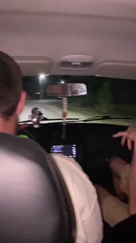 This is a video from inside a car at night. The camera is pointed towards the front of the car and shows the backs of the heads of the people in the front seats. The driver is visible, and in the back, there are two people. One person in the back is facing forward and appears to be a male with short brown hair. Another person in the back is visible from the side and is also male, with short blonde hair and a mustache. He has tattoos on his arm and is looking towards the camera and smiling. The third person in the back is seen from behind, wearing a grey shirt, and appears to be a male. The car is moving, and the outside is dark with some lights visible from street lamps and trees. The people in the car seem to be enjoying themselves and interacting with each other.
