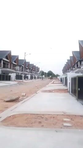 The video shows a street lined with houses under construction. The houses have modern architecture with dark sloping roofs and white walls. The street is paved with concrete, and there is construction debris and dirt on either side. A few streetlights are visible. The sky is overcast. There are no people visible in the video.