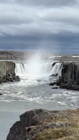 The video captures a majestic waterfall under a heavily overcast sky. The powerful waterfall cascades between dark, rugged cliffs, which appear to be composed of columnar basalt formations. Water plunges into a wide, turbulent river, creating significant mist that rises into the air. The river flows vigorously away from the falls, with white foam and rapids visible. Large, dark rocks are scattered along the riverbanks and in the foreground. The distant landscape beyond the waterfall is rocky and barren, obscured somewhat by the mist and clouds. The scene conveys a sense of raw natural power and grandeur. A voice can be faintly heard in the background. No people or text are explicitly visible.