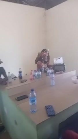 The video shows the interior of a small, sparsely decorated room with light-colored walls and a corrugated metal roof. Two women are sitting behind a wide counter, cluttered with multiple bottles of water, cell phones, and a black bag. One woman is wearing a patterned brown dress and a headwrap. The other woman is partially visible behind her. A man wearing a white shirt and black pants walks into the frame and stands briefly, facing the women before leaving. Another person is seen holding a white object at the right edge of the frame. The scene appears to be a casual gathering or a meeting in a simple setting.