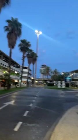 The video pans across an outdoor area, likely an airport or transportation hub, at dusk. Palm trees are prominent in the foreground and middle ground. In the background, a tall, modern building with multiple levels, possibly a parking garage or terminal, is visible. Further back, an air traffic control tower stands against a cloudy twilight sky. Streetlights and building lights illuminate the scene. The ground is paved, with visible lane markings indicating a road. Several concrete benches are placed in a seating area. A white trash receptacle with a polka-dot pattern is also visible. Cars are parked and driving in the background. The overall atmosphere is one of a transition from day to night.