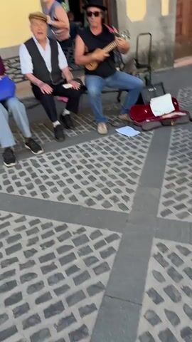 The video opens with a street scene in what appears to be a European town. In the foreground, three men are sitting on a bench or chairs, with one playing a ukulele. A woman is walking behind them, and further back, a man in a yellow shirt is walking towards the camera. The background shows a building with "BBVA" signage, suggesting a bank. The street is paved with cobblestones, and there are buildings lining both sides. As the video progresses, the camera pans to show more of the street, revealing a crowd of people walking and browsing. The architecture is traditional, with balconies and ornate details on the buildings. The scene is bathed in bright sunlight, indicating a clear day. In the distance, a church or cathedral is visible, with mountains in the background. The overall impression is that of a lively and picturesque town square or street, possibly a tourist destination.
