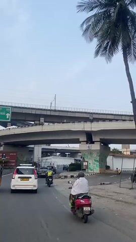 The video shows a view of traffic on a road with multiple elevated flyovers and a train track above. The time is daytime.  On the road, there are several vehicles, including a white car, a scooter, and an auto-rickshaw. A person wearing a white garment is riding a red scooter in the foreground, moving away from the camera. Another person wearing a yellow backpack is riding a motorcycle. The flyovers are made of concrete, and there is some construction or scaffolding visible on one of them. The environment appears to be urban, with some trees and painted murals on the flyover pillars. The sky is overcast.
