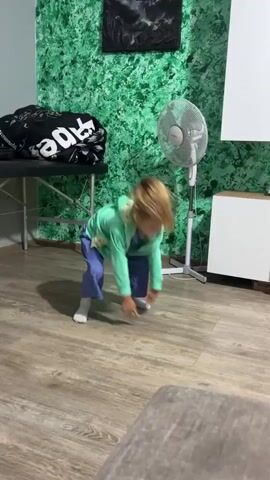 A young girl with blonde hair tied in a ponytail is captured mid-jump in a room with a distinctive green, textured wall. She's wearing a blue jumpsuit and a light green zip-up hoodie, with white socks. Her mouth is open, suggesting she's mid-action or vocalizing. The room appears to be a living space, indicated by the wooden floor and a piece of furniture in the foreground that looks like a sofa. In the background, there's a black and white framed artwork on the wall, a portable massage table covered with a black fabric bearing white text that reads "RIDE", a standing fan, and a white cabinet. The overall lighting suggests it's daytime. The girl's energetic pose and facial expression convey a sense of joy and excitement. The setting seems to be indoors, likely a home environment.