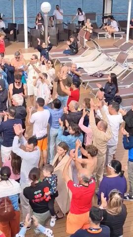 A crowd of people are clapping their hands and moving in a rhythmic fashion on a deck, presumably a ship or large boat, as indicated by the distant sea. The group, comprising adults and children, is dressed in casual attire suitable for warm weather. Various styles of clothing are evident, from t-shirts and shorts to casual pants and dresses. Some individuals are shirtless. Their attention is focused towards something unseen in the video. The flooring of the deck consists of wooden planks, and lounge chairs are arranged along the edges. Additional people can be seen relaxing in the background. Overall, the atmosphere appears festive and celebratory.