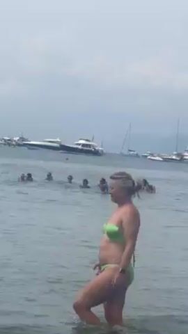 The video shows a group of people swimming in the ocean near several boats.  A woman in a green bikini stands in the foreground, while a group of people are swimming behind her. Some are using floats. There are multiple boats visible on the horizon under a cloudy sky.