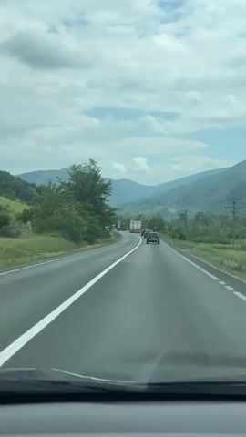 The video shows a first-person perspective from inside a vehicle driving along a two-lane asphalt highway through a scenic, mountainous landscape in Plugova, Romania. The road has clear white lines marking the lanes and shoulder. In the distance, other cars and several large trucks can be seen, suggesting moderate traffic. Lush green trees and vegetation line both sides of the road, and rolling hills and mountains, also covered in greenery, frame the horizon. Power lines with utility poles are visible on the right side of the road. The sky is partly cloudy with patches of blue, indicating a bright, clear day. The overall impression is a peaceful journey through nature. No legible text is visible in the video.