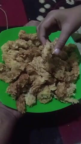 A person's hand is picking up a piece of fried chicken from a bright green plate. The plate is full of fried chicken pieces. The fried chicken appears to be breaded and golden brown, with some pieces darker than others. The background is blurred and dark, with hints of a red and black patterned surface.