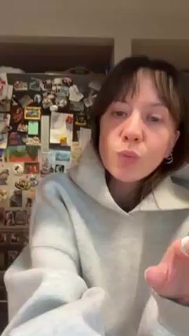 A young woman with medium-length brown hair and bangs is standing indoors wearing a light gray oversized hooded sweatshirt. She starts by waving her hands in front of her face. She then grabs the neckline of her sweatshirt and pulls it away from her neck, shaking her hair out so that it falls down her back. The background shows a refrigerator covered with magnets, photos and notes. A cabinet filled with dishes is on the right. The lighting is warm.