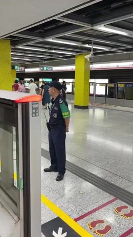 The video shows a train platform. A security guard in a dark uniform with a green armband stands facing left, looking at the train doors. People are walking on the platform. The doors are closed. A man walks past the camera. The camera then moves closer to the train doors, showing reflections of people on the platform.