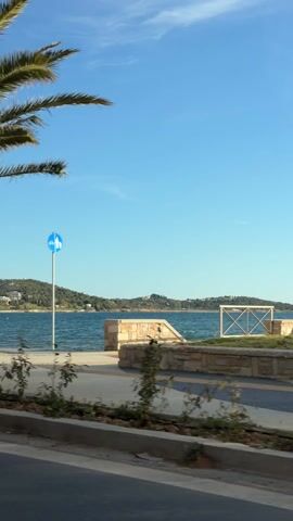 The camera, likely mounted on a moving vehicle, captures a scenic view of a coastline on a bright, sunny day. The foreground consists of a road, followed by a divider with small bushes. Beyond that is a wide promenade with a red walking path, separated from the water by a low white wall. The water itself is a deep blue with gentle waves. In the distance, there's a green, hilly landmass dotted with white buildings, suggesting a residential or resort area. The sky is a clear, light blue with a few wispy clouds. Occasional trees and palm fronds pass by in the foreground, obscuring the view momentarily as the vehicle moves. The overall impression is one of a pleasant coastal environment.