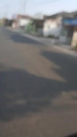 The video is a first-person perspective video of a road with buildings on the side. The video is blurred, making it difficult to discern specific details. There are a few blurry figures that appear to be people walking on the sidewalk. There are also a few blurry vehicles on the road. The sky is clear and blue. The sun appears to be shining. The overall impression is of a quiet, residential street.
