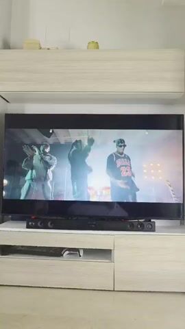 A TV screen displays a music video. Three men are initially seen on stage, one wearing a Bulls jersey. The next shot shows a group of people raising their hands, including some holding red plastic cups. A sequence of shots follow, showing several women dancing and posing for the camera. The TV is placed on a white cabinet unit in what appears to be a living room. Below the TV is a sound bar and a PlayStation console in a drawer.