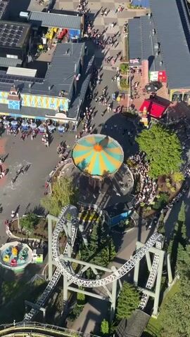 This is an aerial drone shot of an amusement park. The camera is moving downwards and slightly to the right, showing a carousel with a green and orange striped roof in the center of the frame. A roller coaster track is visible in the foreground, with a few coaster cars on it. There are many people walking around the park, some in lines for rides. Buildings with shops and attractions are visible. Green trees and landscaping are scattered throughout the park. The overall scene depicts a busy and lively amusement park on a sunny day.