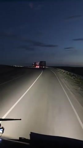 The video shows a view from inside a vehicle traveling on a highway at dusk. The sky is a deep blue with scattered clouds, indicating either the beginning of the night or the end of the day. The headlights of the vehicle illuminate the road ahead, which curves to the right. In the distance, there are lights from what appears to be a settlement or town. Two large trucks are visible on the road ahead, their taillights glowing red. The landscape appears to be flat and barren, with some scrubby vegetation visible on the side of the road. The overall mood is one of solitary travel through a vast, open landscape as darkness descends. No visible text is present in the video. The context provided indicates the location is Baiqadam, Kazakhstan.