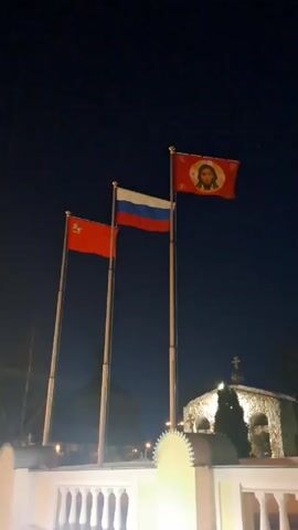 At night, three flags stand in front of a small, stone building with a cross on top. The flag on the left is red, the one in the middle is the Russian flag, and the one on the right is red with an icon of Jesus on it. They are all waving gently in the breeze against a dark sky. The scene is lit by several unseen light sources.