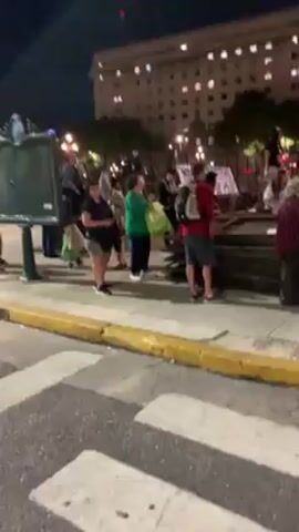 The video depicts a night scene in what appears to be a city square. A small group of people is gathered near a metal barricade and a dark green information stand. The people are dressed in casual clothing and some are carrying bags. In the background, there is a large building illuminated by lights. A statue is also visible on the left. The scene is shot from across a crosswalk.