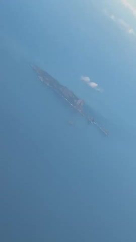 An aerial view from an airplane window shows a long, narrow island surrounded by a vast blue ocean under a clear blue sky. A small white cloud is visible in the distance near the island. As the video progresses, the aircraft's white wing and engine become visible in the foreground, framing the distant island and ocean.