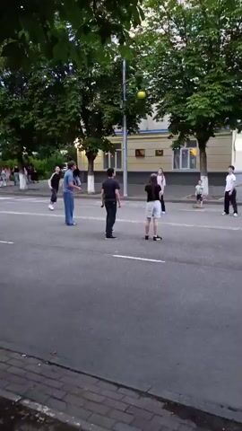 A group of young people are playing volleyball in the street. A yellow ball is in the air, and one person is jumping to hit it. The other people are watching or preparing to hit the ball. There are trees and a building in the background.