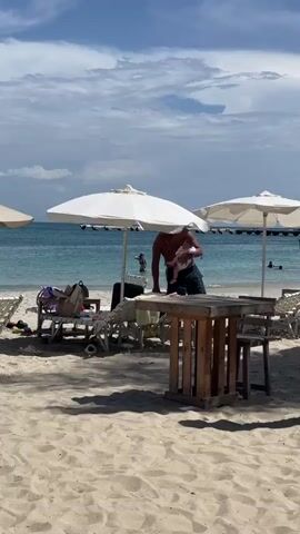 The video shows a beach scene. In the foreground, there is sand and a wooden table with chairs. Behind the table, there are lounge chairs and umbrellas. Further back, people are in the ocean, some swimming and some standing in the water. The sky is blue with scattered clouds.