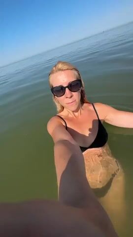 A blonde woman wearing sunglasses and a black bikini swims in calm, greenish water. She is holding a phone or camera out in front of her, taking a selfie. Sunlight reflects off the water.