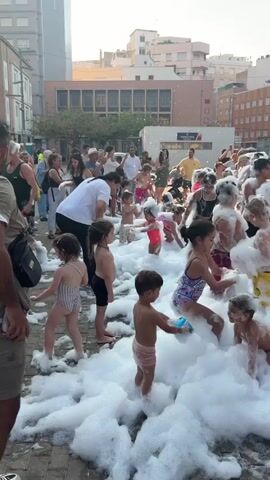 The video shows a lively outdoor foam party, likely in a town square, filled with many children and some adults submerged in a thick layer of white foam. The atmosphere appears festive and energetic, with people of all ages interacting and playing in the foam. Most of the children are wearing swimsuits, indicating a water-related activity. Some adults are also participating, either by supervising or joining in the fun. The background reveals buildings typical of a European urban setting, with some trees and a bright sky suggesting it is daytime. The foam is being generated from a machine visible in the background. The overall scene captures a joyful community event.
