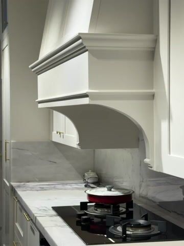 The video shows a white kitchen with marble countertops and backsplash. A red pot sits on a black stovetop beneath a decorative white range hood. The cabinets are white with gold handles.