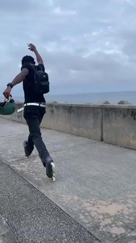 A person wearing black pants, a black t-shirt, and a black backpack is inline skating along a paved walkway next to a concrete barrier. The person is holding a green helmet in their right hand and a phone in their left hand. The sky is cloudy, and there is a body of water visible in the background. The person is moving from left to right in the frame, with their left leg extended forward. The wheels of the inline skates are visible and appear to be spinning. The person's body is slightly contorted as they are in motion.