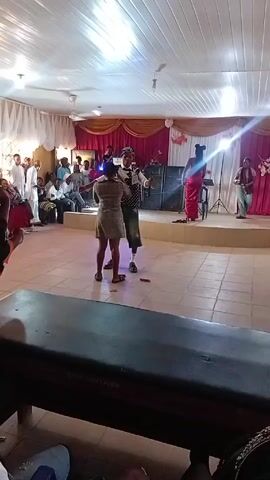 Here's a description of the video:

The video shows a group of people gathered inside a large room for what appears to be a celebration or event. A stage is set up at one end of the room, decorated with pink and gold draping. Several people are seated in rows of chairs, watching the performers.

On the stage, there are at least four people. One is singing into a microphone, and another is playing a guitar. In front of the stage, a man wearing a vintage-style outfit is dancing with a woman in a short dress. Another woman, wearing a long red garment, is also dancing.

The room has a tiled floor and is well-lit with overhead lighting. Some people in the audience are holding up their phones, likely recording the performance. A long dark table sits in the foreground of the shot.